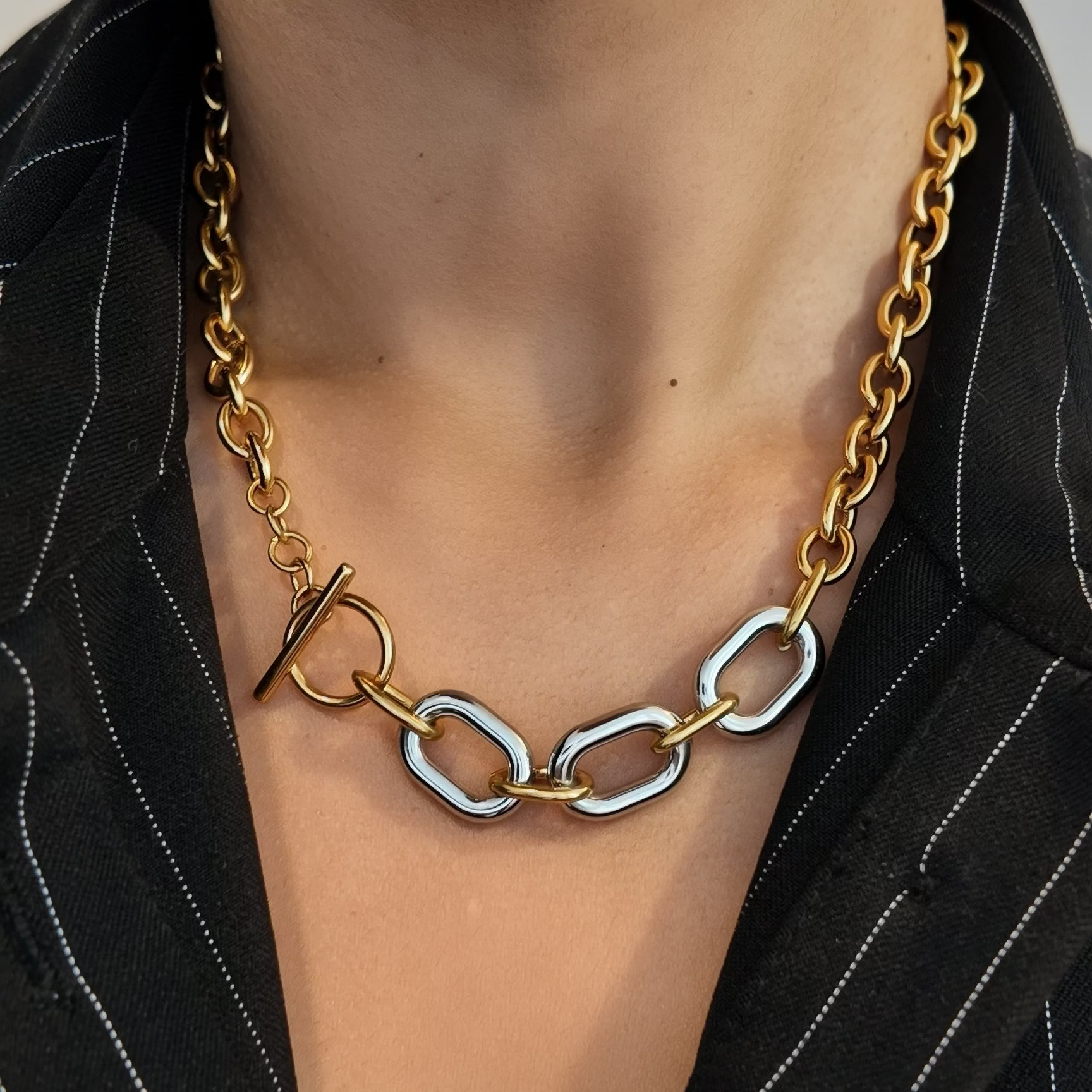A close-up of a person wearing a chunky gold chain necklace with a toggle clasp and three large interlocking silver oval links, styled with a black pinstripe blazer.