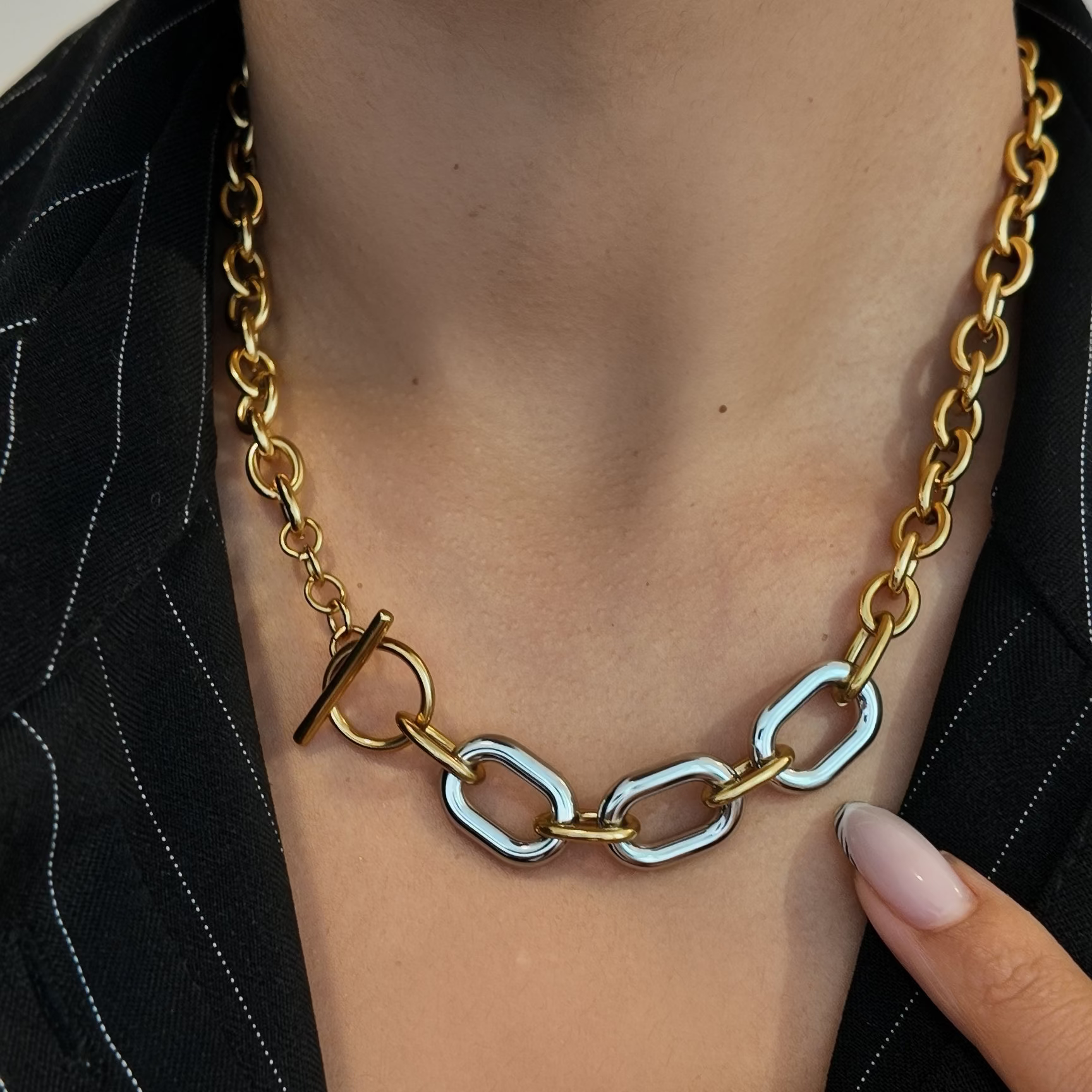 A close-up of a model wearing a gold chain necklace with a unique link design featuring one large gold link and two smaller silver-toned oval links, styled with a black pinstripe shirt.