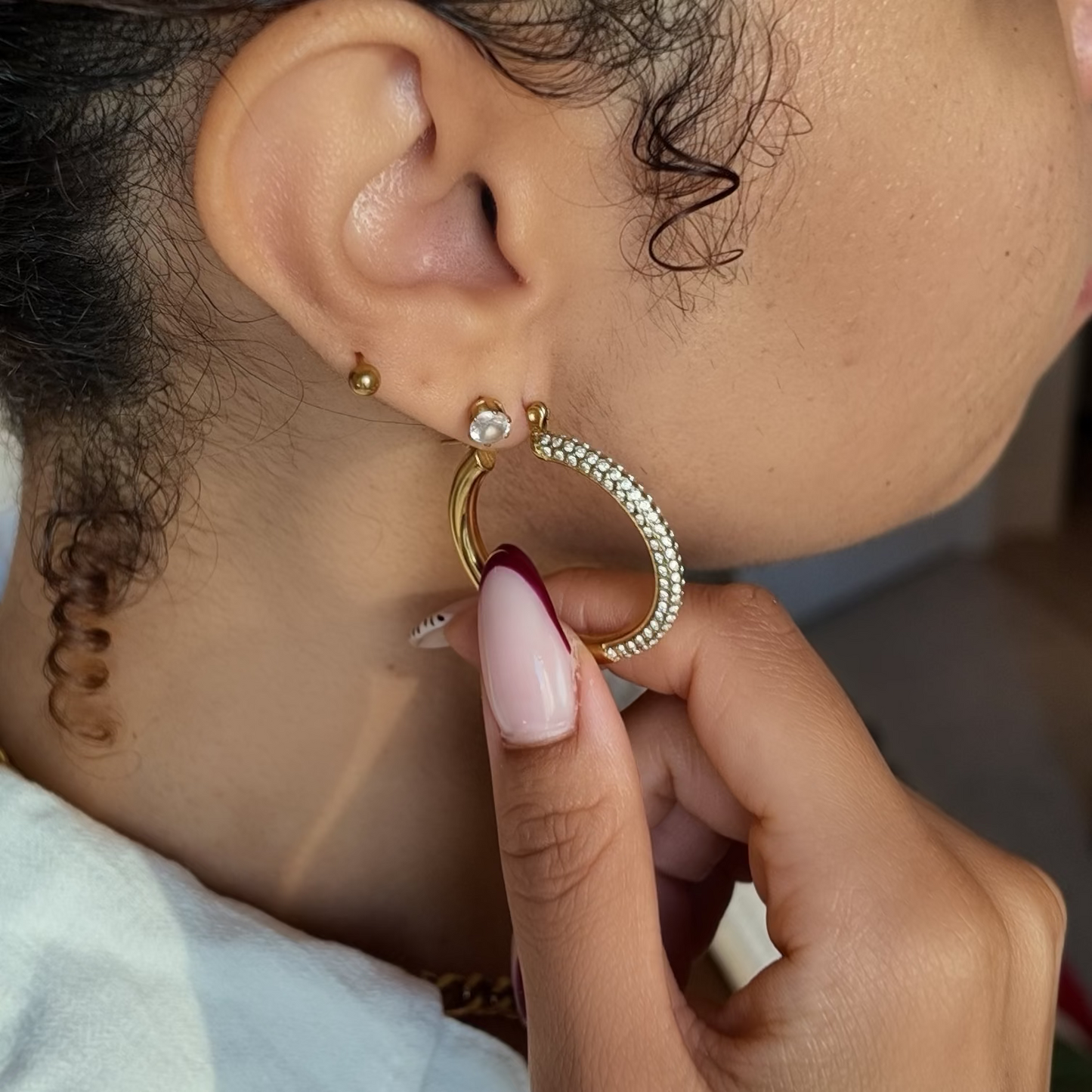 A close-up lifestyle shot of a woman wearing a gold hoop earring adorned with small clear crystals, with her hand gently touching the earring.