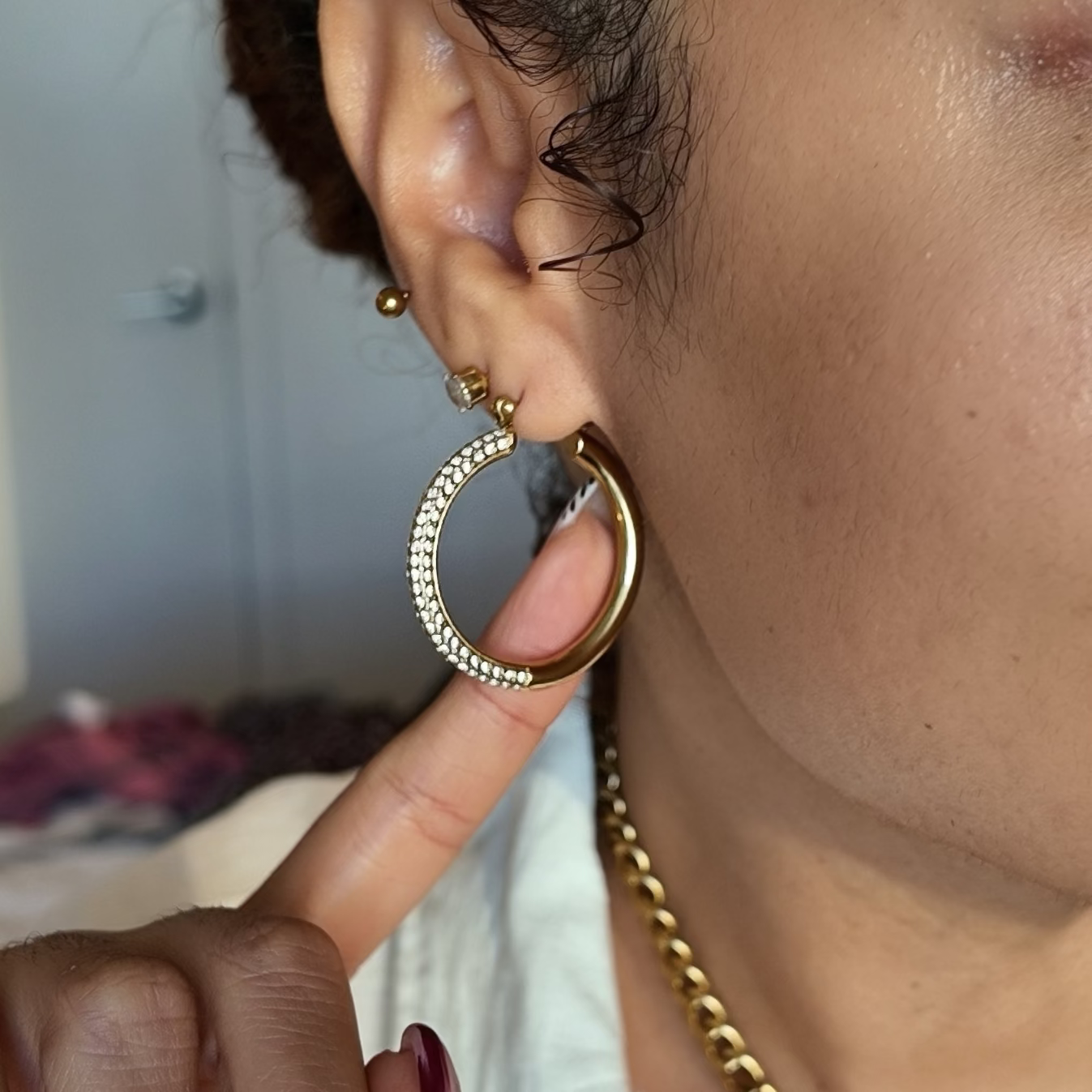 A close-up lifestyle shot of a woman wearing a large gold hoop earring adorned with small clear crystals, with her finger pointing toward the earring.