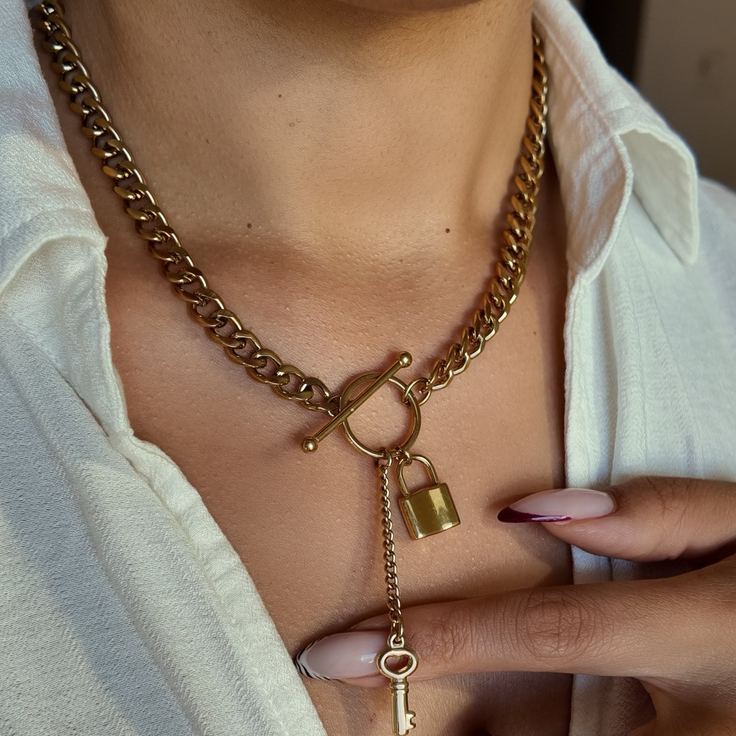 A close-up of a person wearing a gold-toned chain necklace with a toggle clasp, a small padlock charm, and a dangling key pendant, styled with an open white shirt.