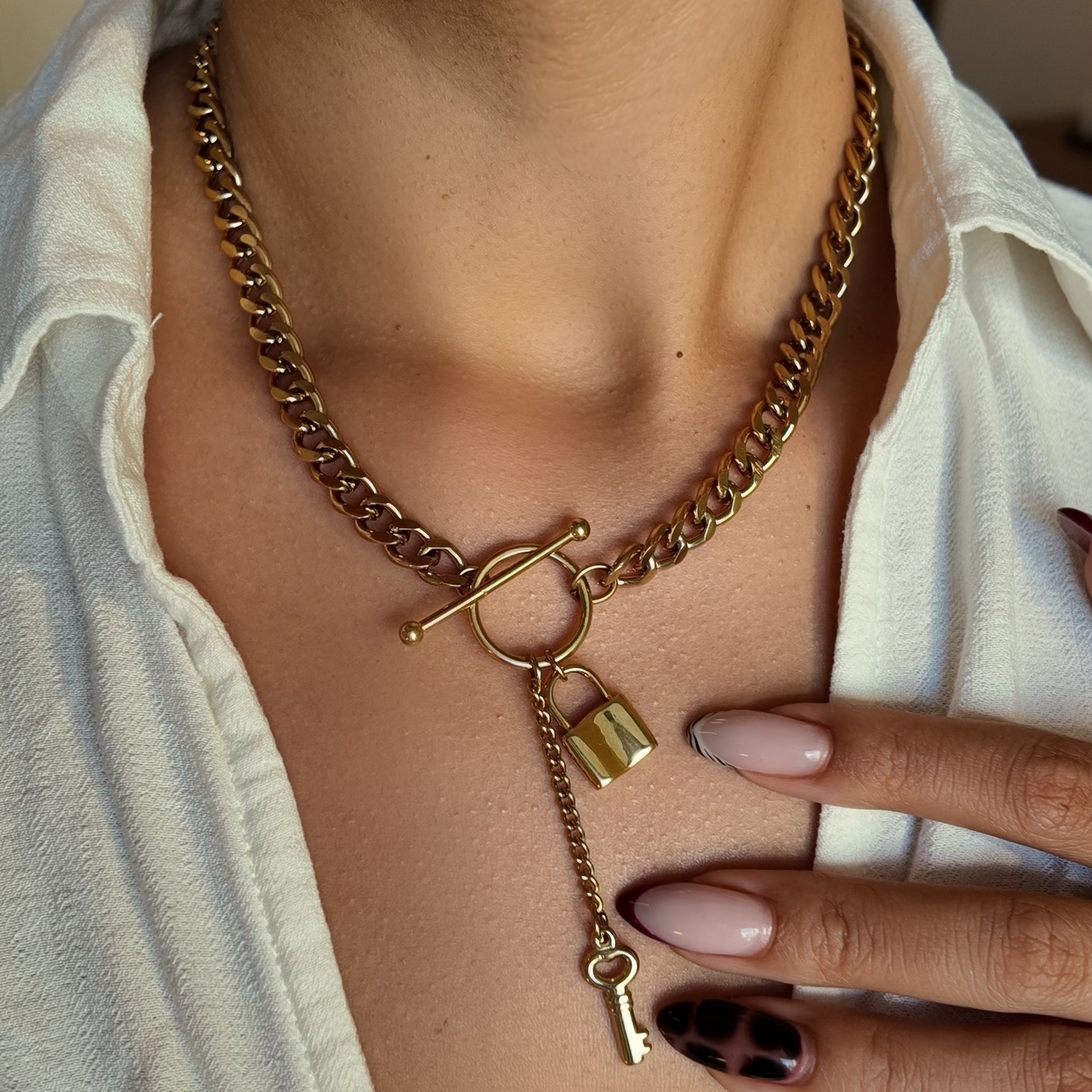 A close-up of a person wearing a gold-toned chain necklace with a toggle clasp, a small padlock charm, and a dangling key charm, paired with a white collared shirt.