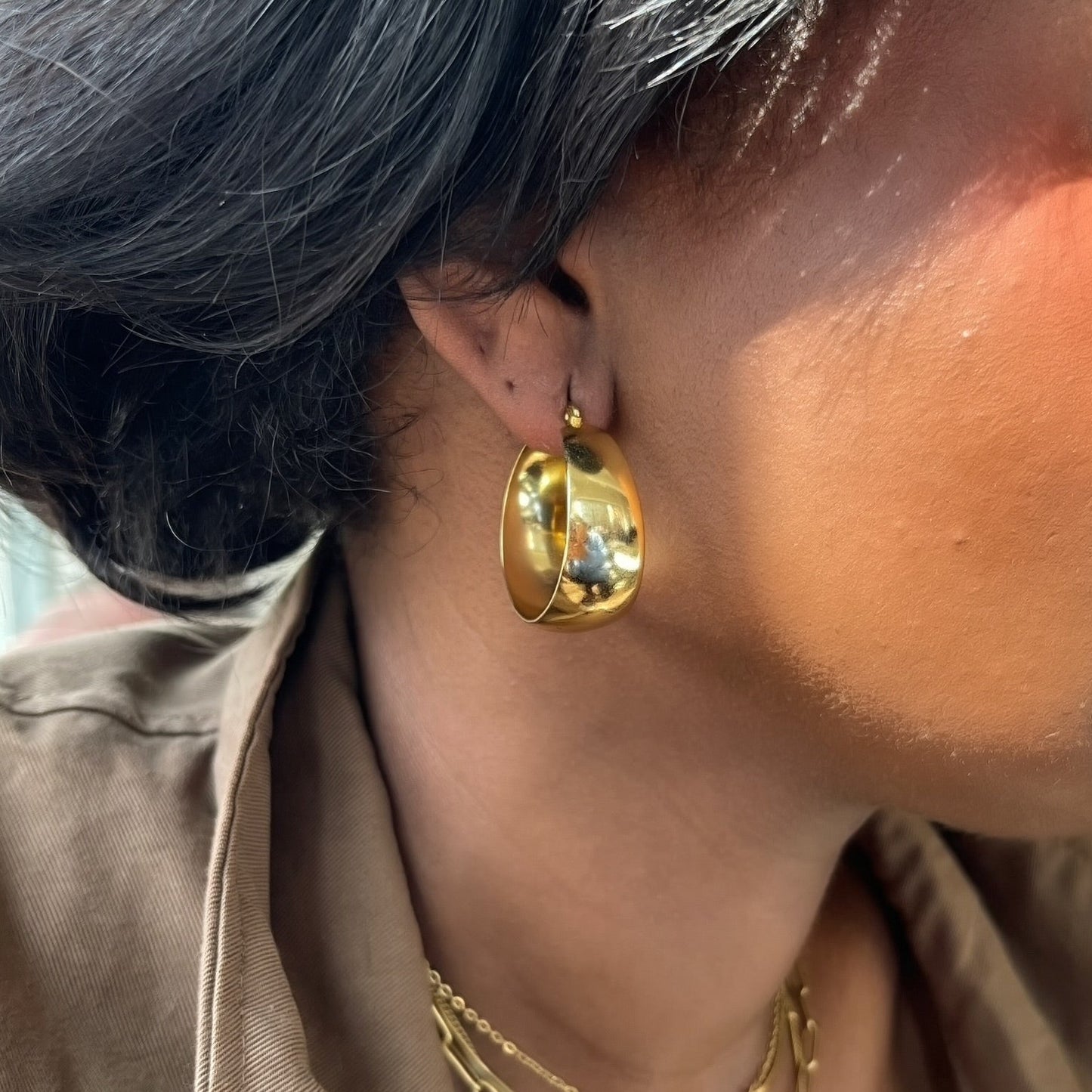 Close-up of a person wearing gold hoop earrings and necklaces with a neutral background.