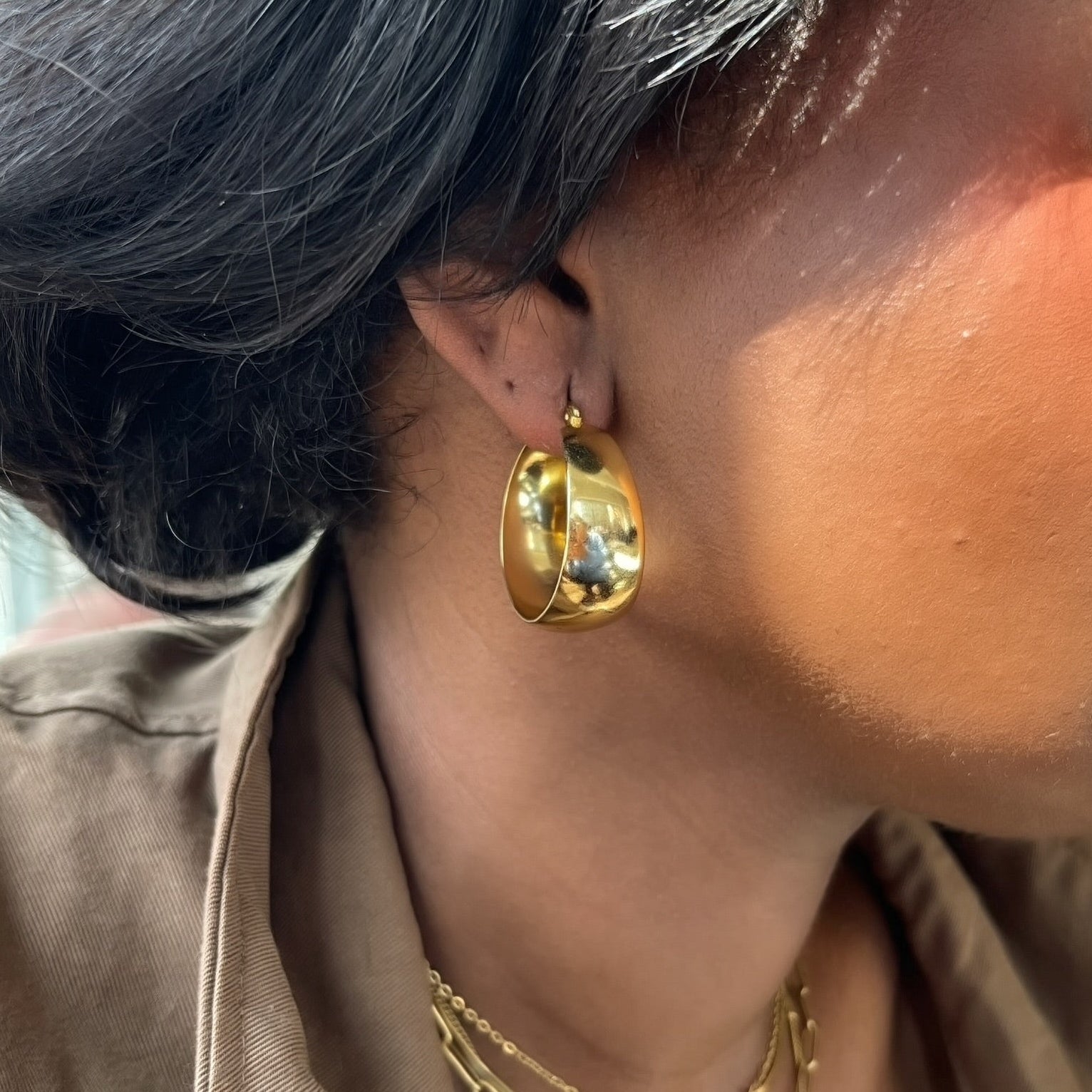 Close-up of a person wearing gold hoop earrings and necklaces with a neutral background.