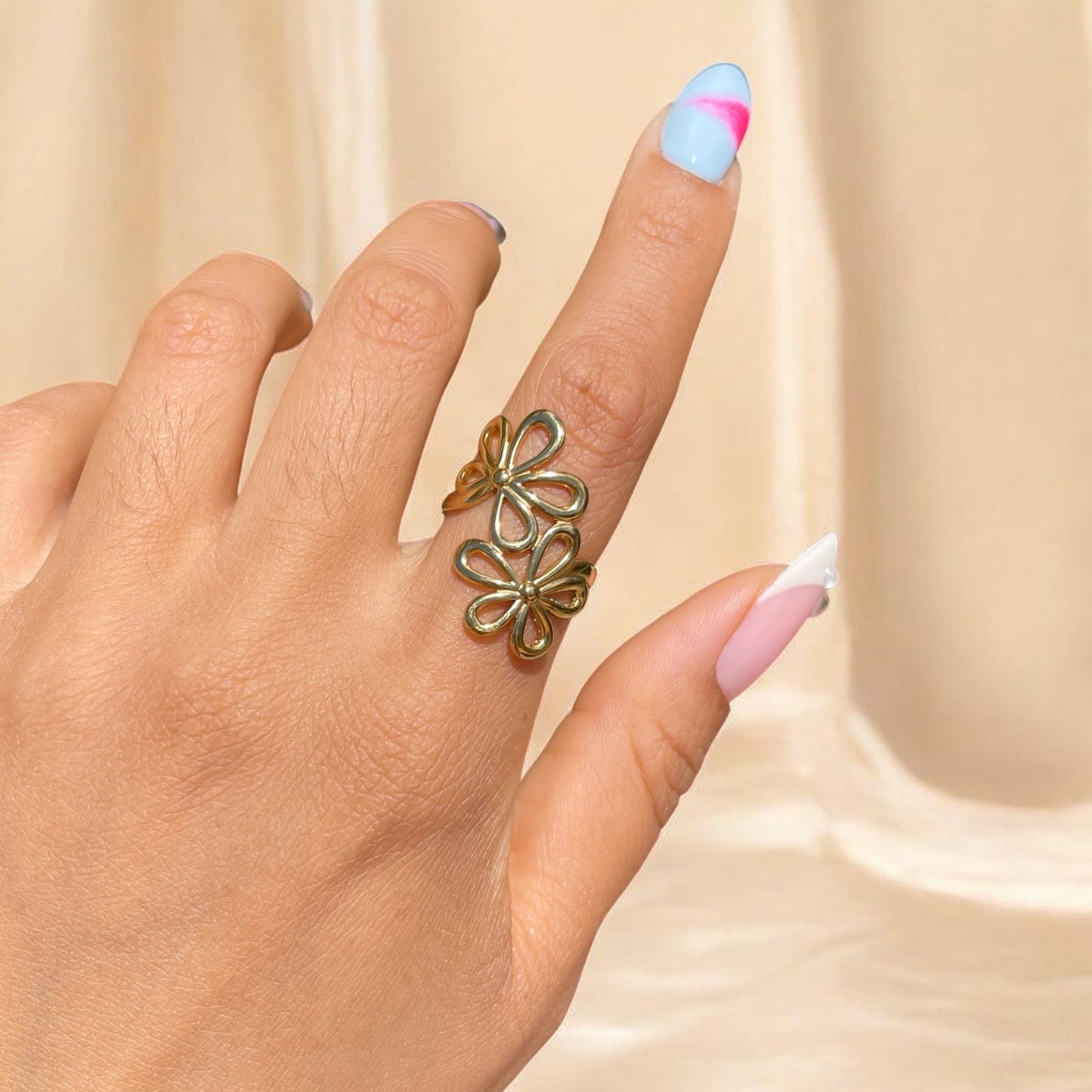 Hand wearing a gold floral ring with pastel nail polish on a beige background