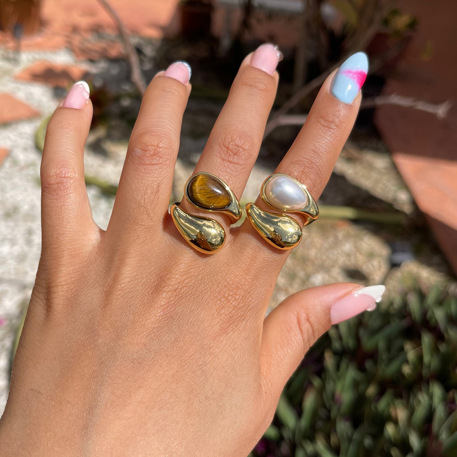 Hand wearing a gold ring with a natural stone, set against a blurred outdoor background.
