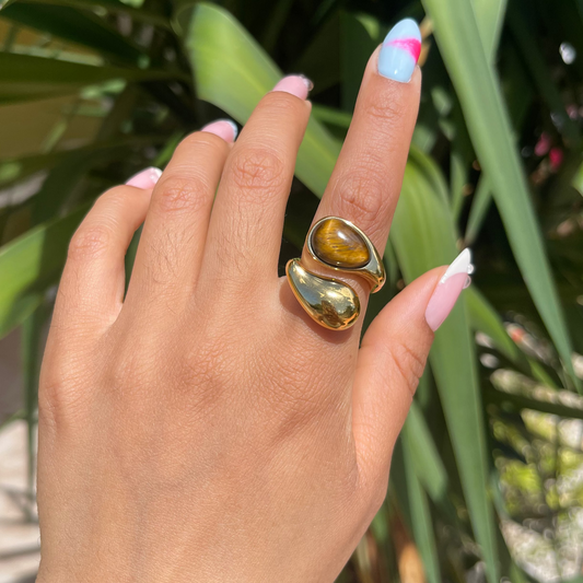 Hand wearing a gold ring with a large gemstone against a green leafy background