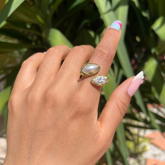 Hand wearing two gold rings with large gemstones against a green leafy background