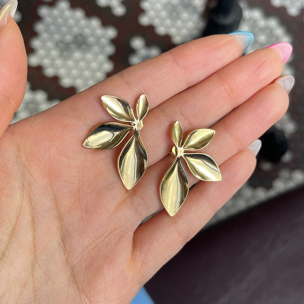 Gold leaf-shaped earrings held in a hand with a patterned background