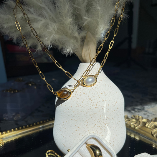Gold necklace with pearls on a white vase with feathers, surrounded by gold jewelry on a dark surface.