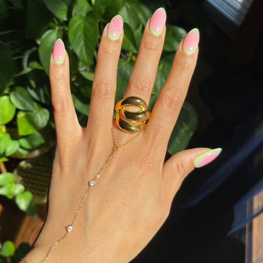 Hand wearing a gold ring with a large gemstone, set against a dark background with greenery.