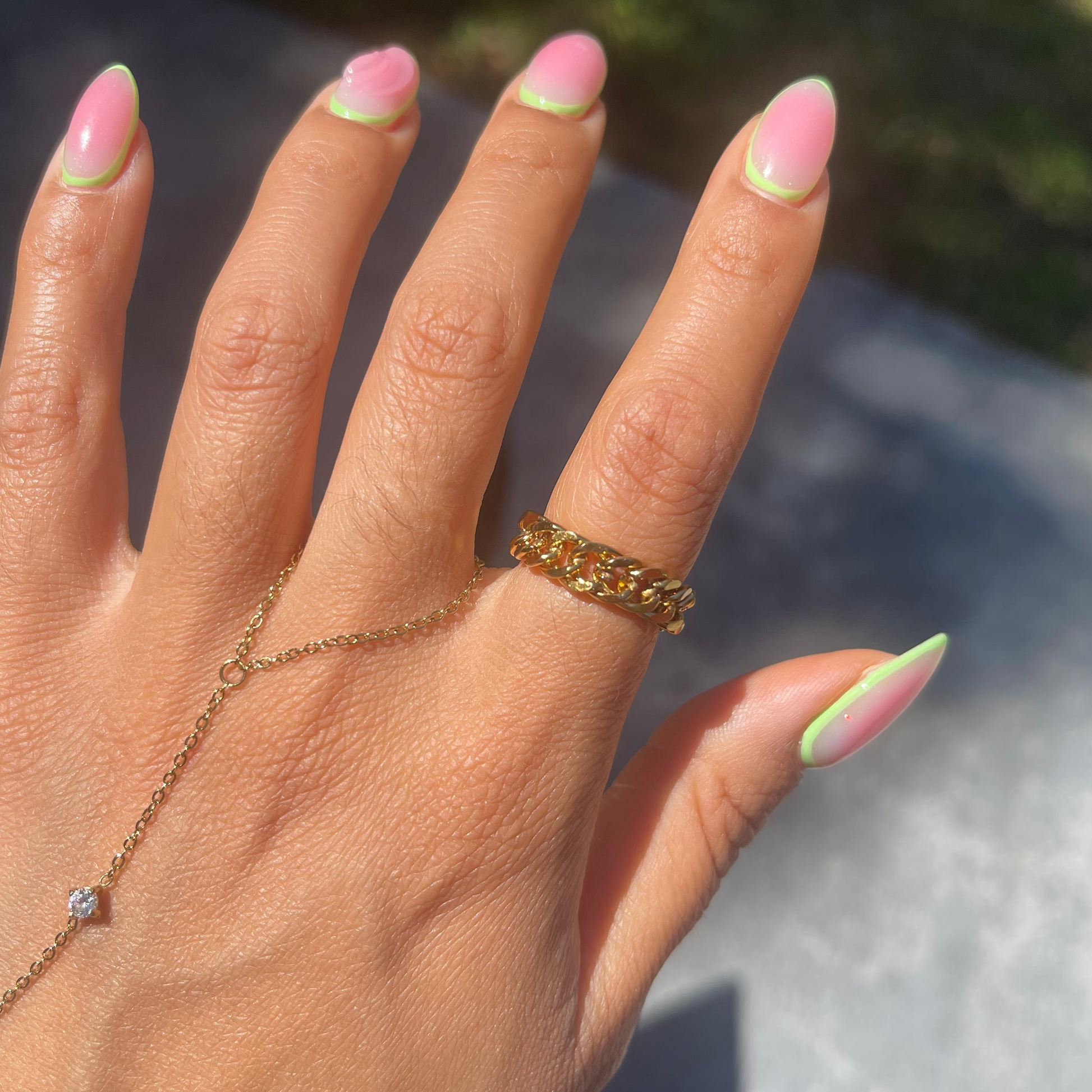 Hand with pink and green nail polish wearing a gold ring and bracelet outdoors.