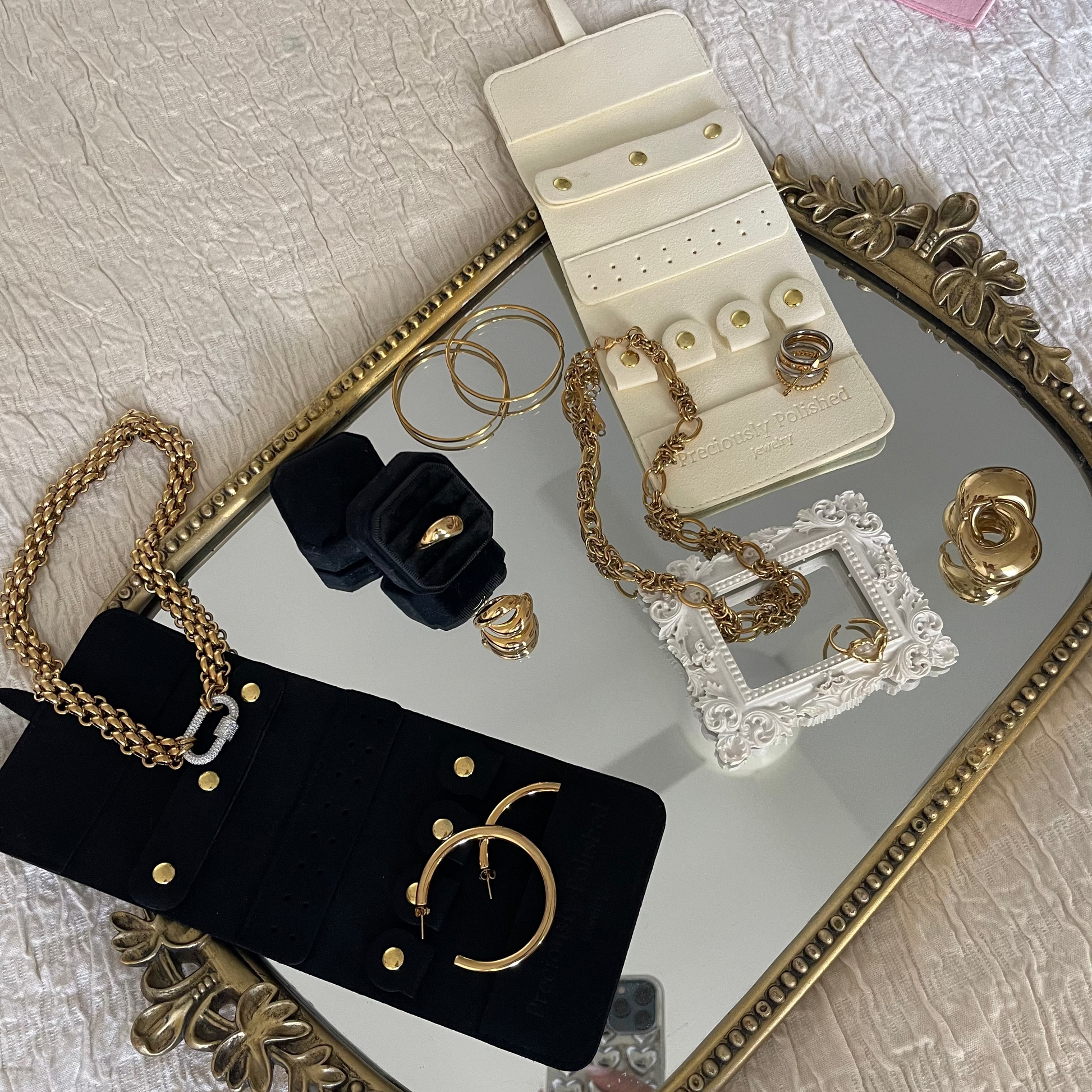 A styled flat-lay of jewelry accessories on a decorative gold tray over a white textured fabric. Visible items include a black velvet jewelry pouch with gold studs and chain, a cream ring display stand, gold rings and chains, a small pink pouch, and a white lace frame.