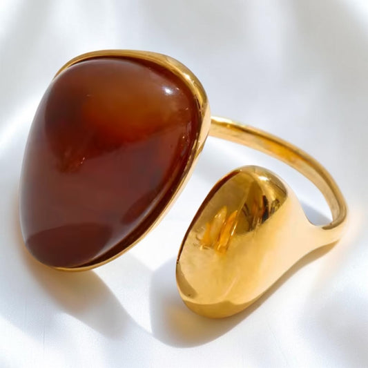 A gold ring with a large, smooth, teardrop-shaped dark brown cabochon gemstone, photographed on a white fabric background.