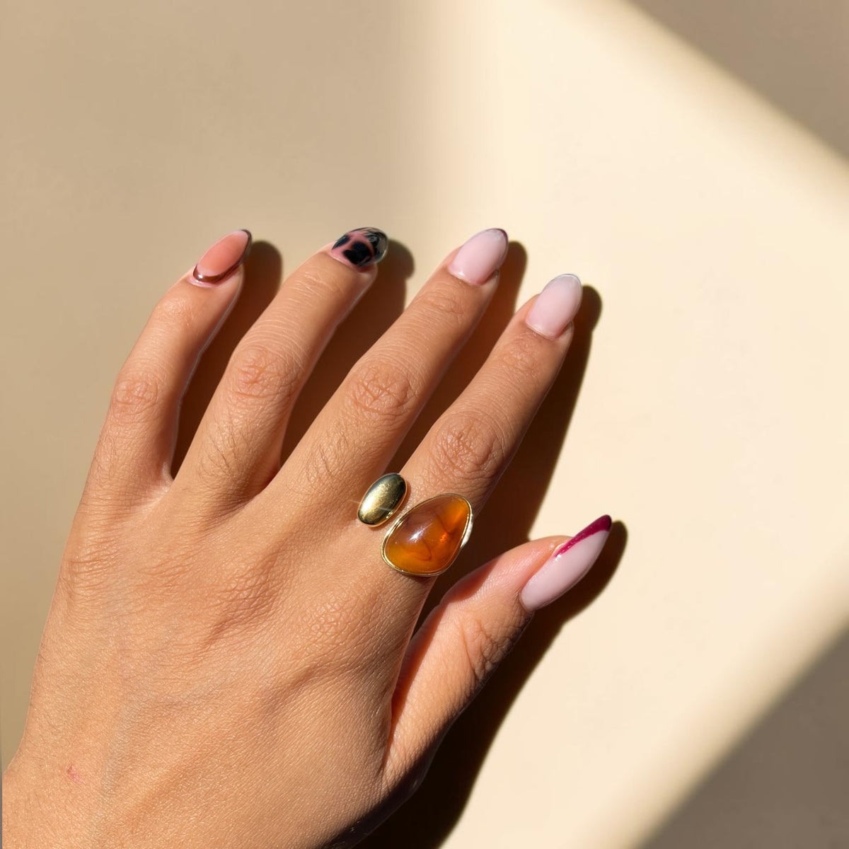 A close-up of a hand with manicured nails wearing two rings: a gold band with a small oval green stone and a larger silver-toned ring with a teardrop-shaped amber cabochon, set against a warm beige background with soft diagonal shadows.
