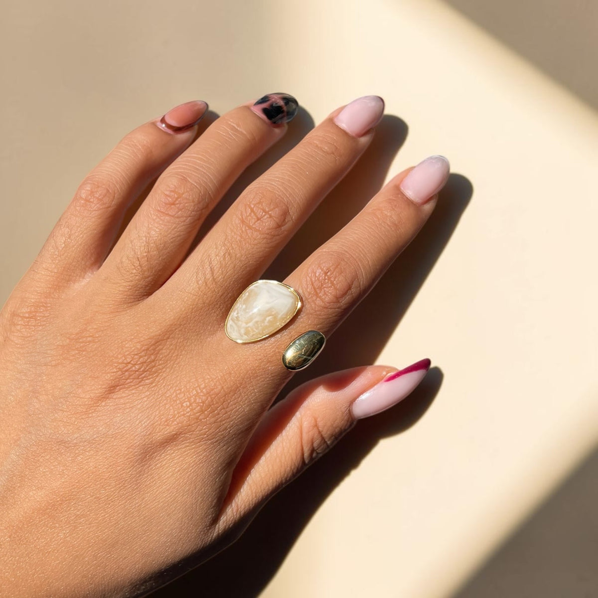 A close-up of a hand wearing two rings: a larger pear-shaped cream-colored stone ring on the middle finger and a smaller oval green stone ring on the ring finger, set against a warm beige background with strong sunlight and shadows.