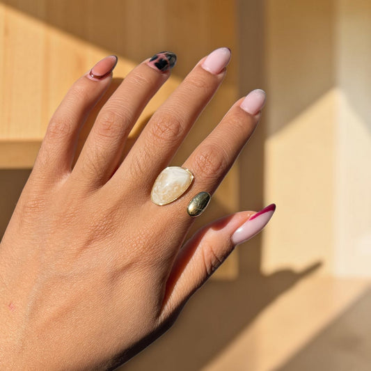 A close-up of a hand wearing two rings: a large cream-colored stone ring and a smaller metallic gold ring, set against a warm beige background with soft shadows.