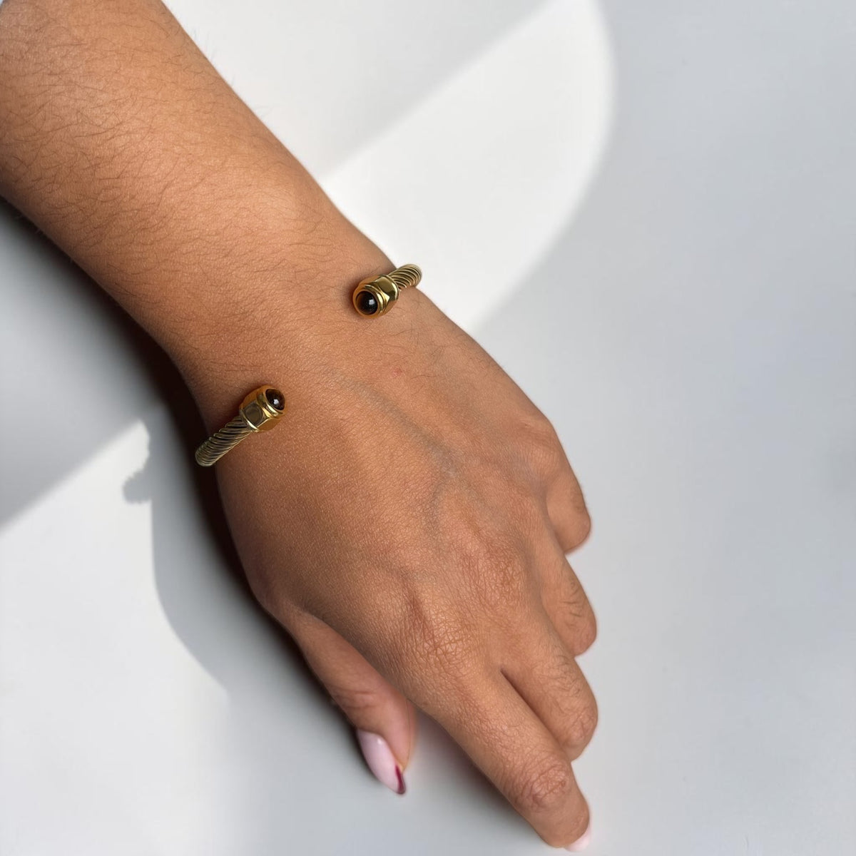 A close-up of a person's hand and wrist wearing two gold-toned cable-style cuff bracelets with small round bead details, set against a clean white background.