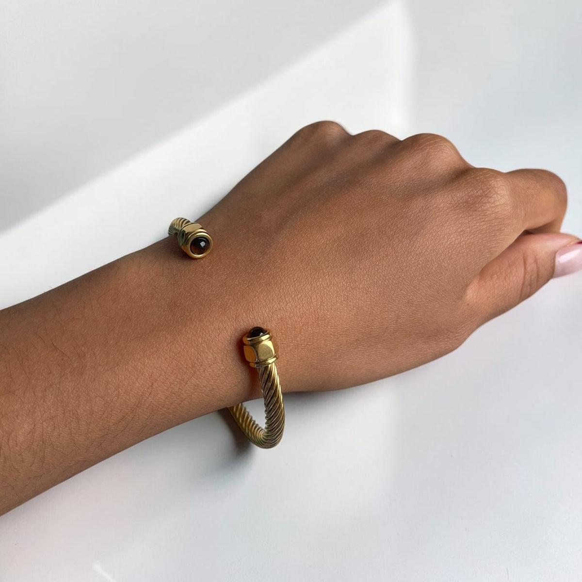 A close-up of a person's wrist wearing two gold-toned bracelets on a clean white surface. One bracelet is a slim bangle with a dark round stone, and the other is a thicker rope-style bangle with a matching dark stone.