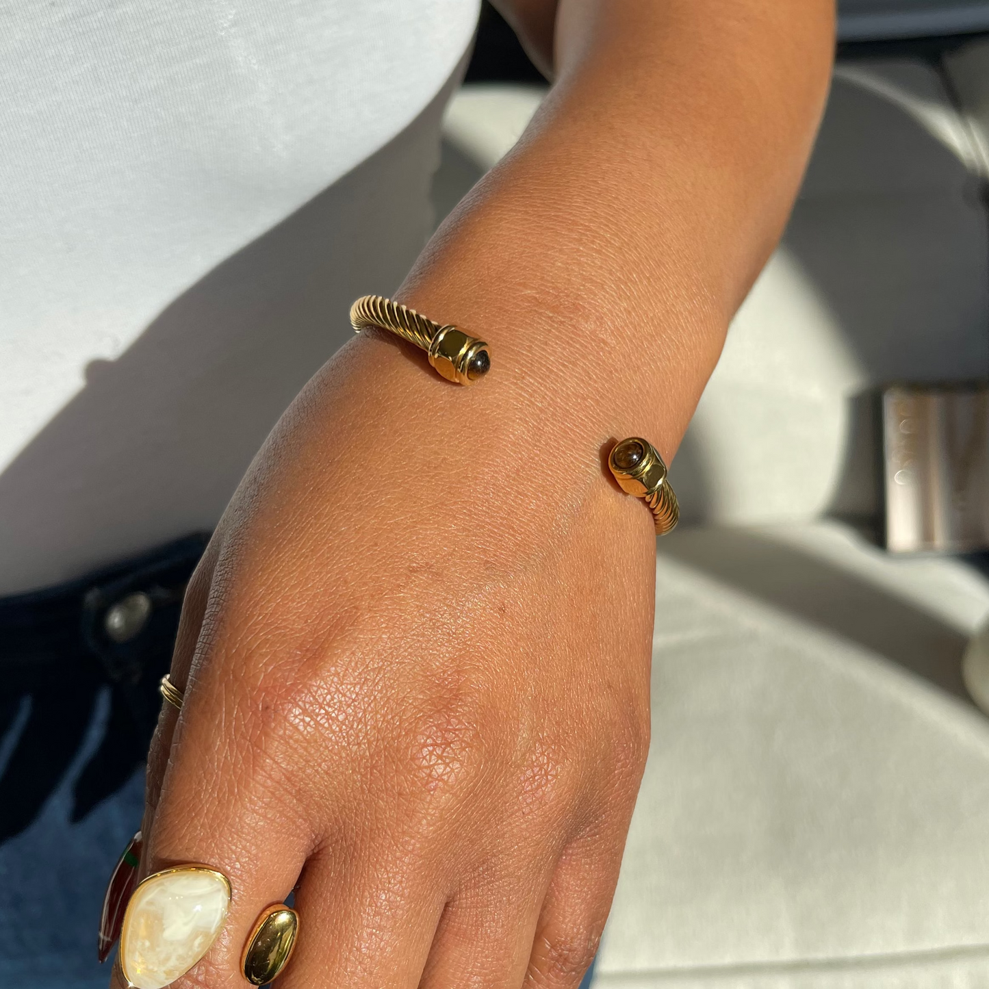 A close-up of a person's hand and wrist wearing two gold-tone bracelets, one textured rope-style bangle and one slim polished bangle, paired with a white top and blue jeans.