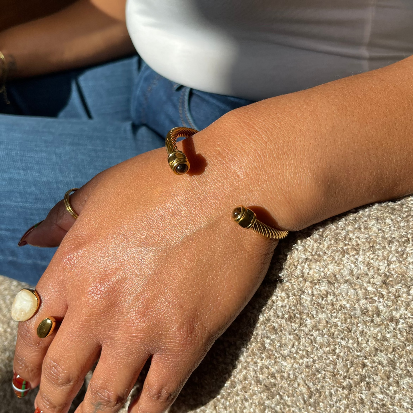 A close-up lifestyle shot of a person's hand resting on a textured beige sofa, wearing two gold snake cuff bracelets and a gold ring with a white stone.