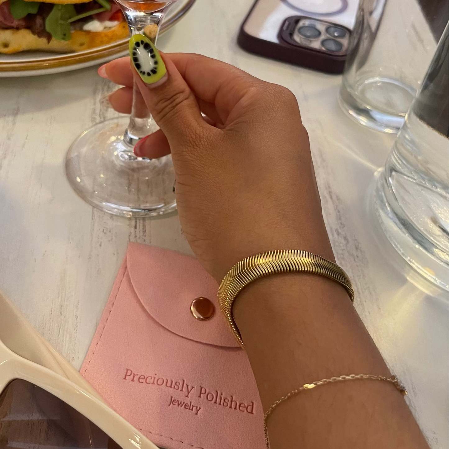 Person holding a cocktail glass with a drink, wearing a thick herringbone bracelet surrounded by a table setting with food and accessories.