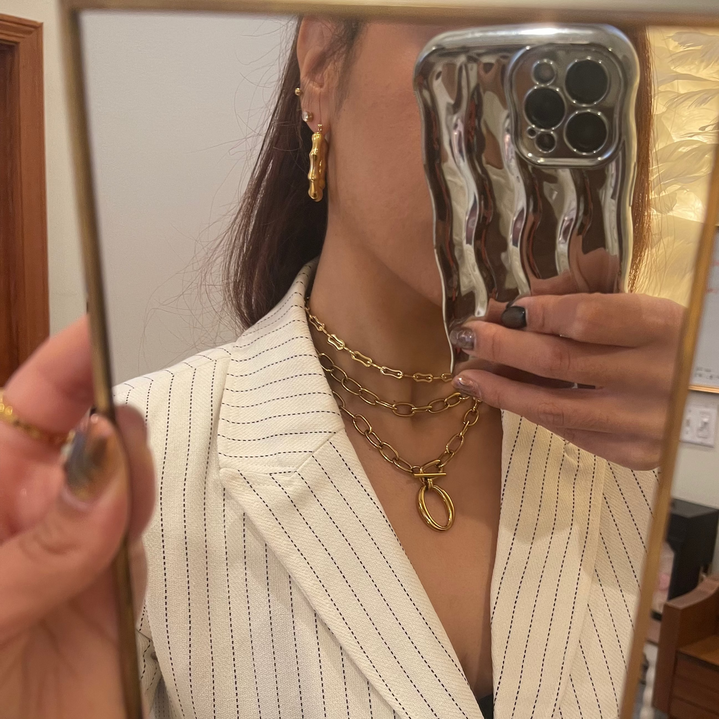 Person holding a reflective phone case in front of a mirror, wearing a white blazer and gold jewelry.