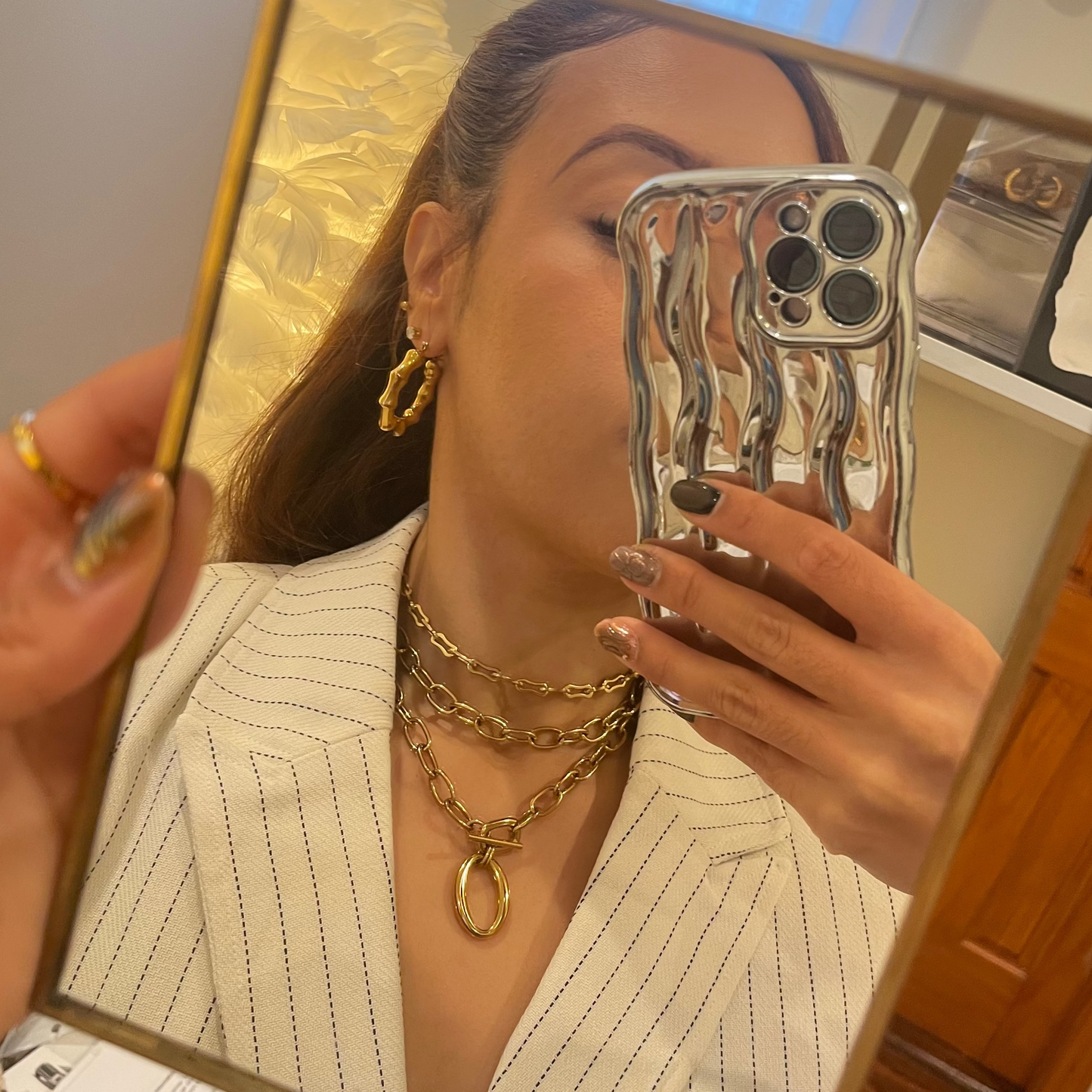 Person wearing gold jewelry taking a mirror selfie with a phone case featuring a wave design.