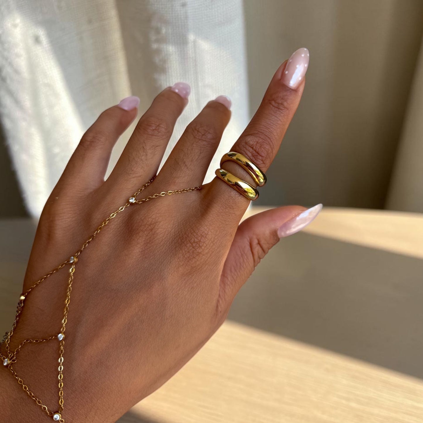 A close-up of a hand wearing a gold chain hand bracelet and two stacked gold rings, set against a soft-focus background of light curtains and a wooden surface.