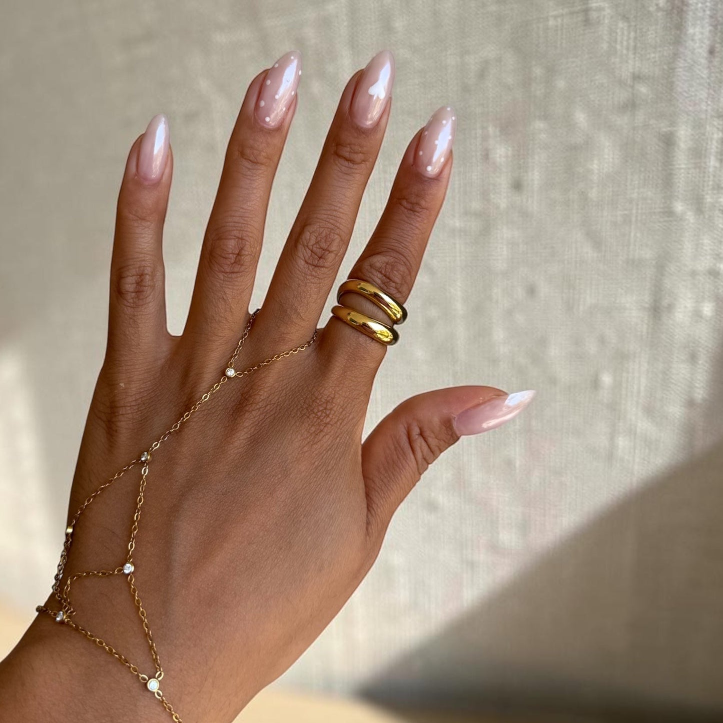 A close-up of a hand wearing a delicate gold chain hand bracelet, matching gold chain wrist bracelet, and two stacked gold rings, set against a textured neutral background.